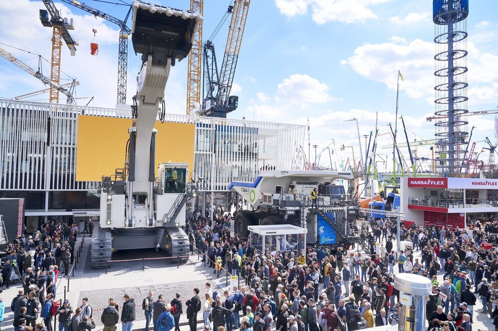 Eine große Menschenmenge auf einer Baumaschinenausstellung besichtigt unter einem teilweise bewölkten Himmel riesige Maschinen und Kräne.