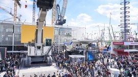 Eine große Menschenmenge auf einer Baumaschinenausstellung besichtigt unter einem teilweise bewölkten Himmel riesige Maschinen und Kräne.