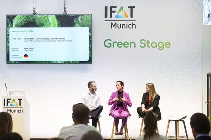 Three people are sitting on the stage of the Green Stage at IFAT Munich, data on the presentation is displayed on a screen.