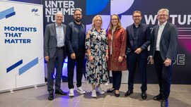 Vier Männer und zwei Frauen posieren gemeinsam für ein Gruppenbild vor einer bunten Messeleinwand und neben dem Motto „MOMENTS THAT MATTER“.