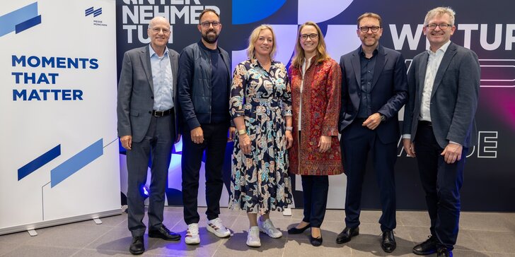 Vier Männer und zwei Frauen posieren gemeinsam für ein Gruppenbild vor einer bunten Messeleinwand und neben dem Motto „MOMENTS THAT MATTER“.