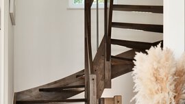 Moderne Holztreppe mit offenen Setzstufen, daneben eine schwarze Vase mit Pampasgras in einem hellen Korridor.