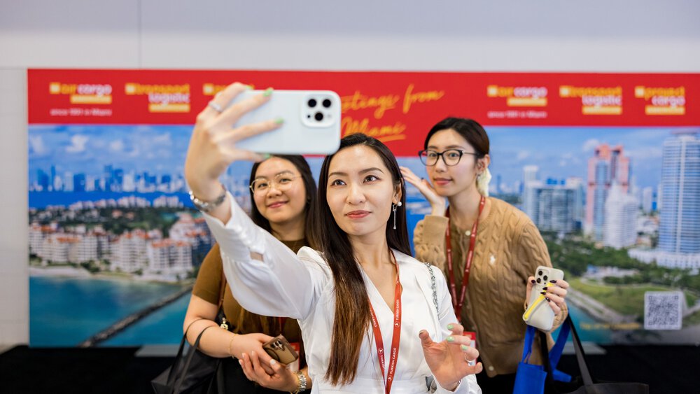 Drei Messebesucherinnen lächeln gemeinsam für ein Selfie vor einer Fotowand der transport logistic Messe.