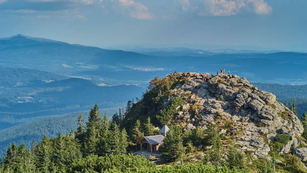 Auf einem felsigen Gipfel stehen Menschen und blicken über den Wald und in der Ferne auf blaue Hügel unter einem wolkigen Himmel.