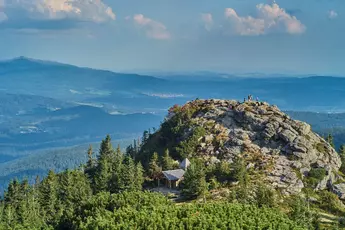 People stand on a rocky peak and look out over the forest and blue hills in the distance under a cloudy sky.