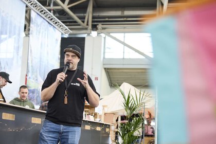 Ein Mann mit schwarzem Shirt und Kappe spricht mit einem Mikrofon auf einer Messe, während im Hintergrund Stände und Zelte zu sehen sind.