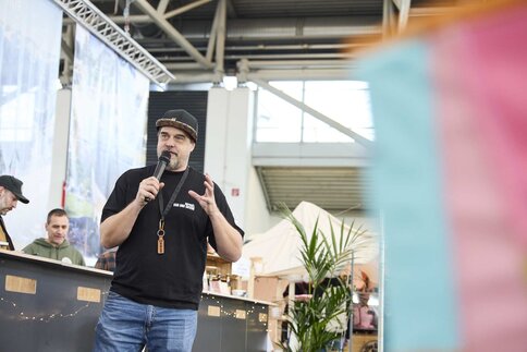 Ein Mann mit schwarzem Shirt und Kappe spricht mit einem Mikrofon auf einer Messe, während im Hintergrund Stände und Zelte zu sehen sind.
