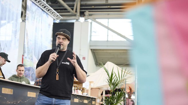 Ein Mann mit schwarzem Shirt und Kappe spricht mit einem Mikrofon auf einer Messe, während im Hintergrund Stände und Zelte zu sehen sind.