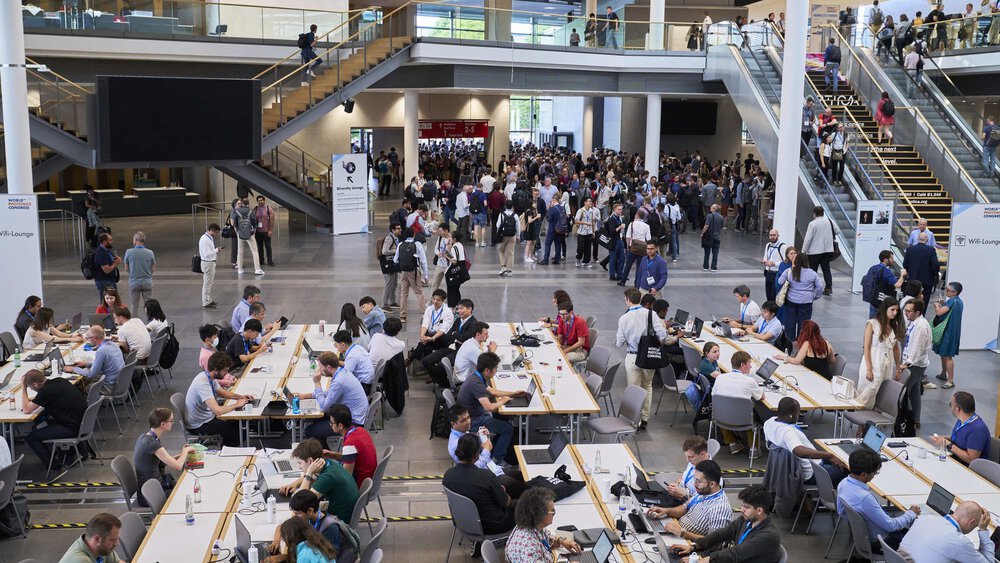 An area with tables and chairs within a large exhibition hall, where people are sitting with their laptops and cell phones.