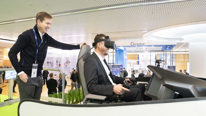 Ein Mann sitzt in einem Simulator an einem Messestand und trägt ein VR-Headset, ein weiterer Mann sieht ihm über die Schultern, beide lachen.