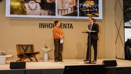 Two people are speaking on stage at a jewellery event, while images are displayed on a large screen behind them.