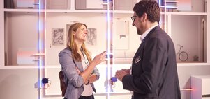 Eine Frau und ein Mann in Geschäftskleidung unterhalten sich lachend. An der Wand hinter ihnen ist ein Heizungssytem am Querschnitt eines Gebäudes illustriert.
