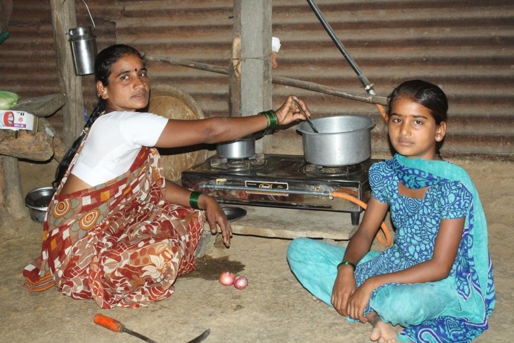 An Indian woman and her daughter are sitting in a wooden hut on a clay floor in front of a gas stove with a pot on top.