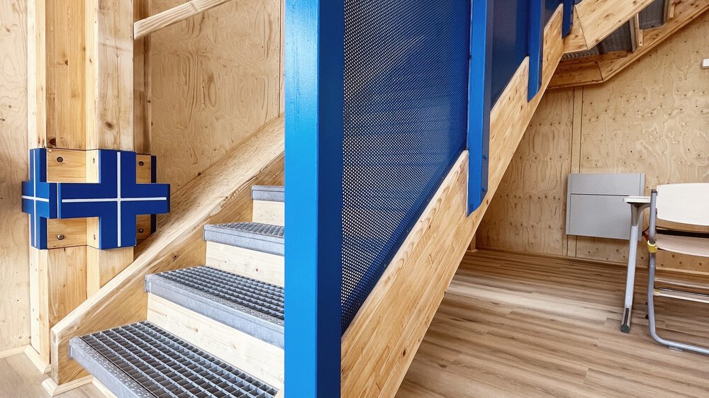 Eine an der Wand entlang nach oben führende Holztreppe mit blauem Geländer. Die Stufen bestehen aus Metallgittern.