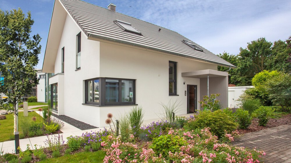 Modernes weißes Haus mit Schrägdach, umgeben von einem üppigen Garten und einer Auffahrt unter klarem Himmel.