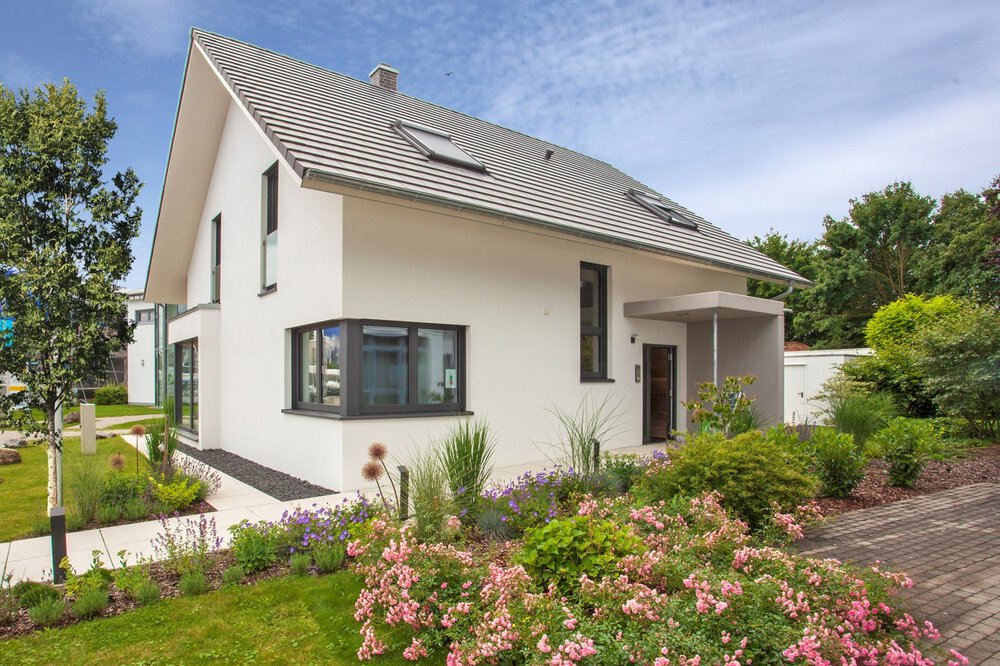 Modernes weißes Haus mit Schrägdach, umgeben von einem üppigen Garten und einer Auffahrt unter klarem Himmel.