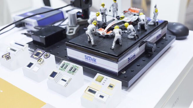 An einem hell beleuchteten Messestand findet in einem Miniaturdiorama ein Reifenwechsel an einem Formel 1 Wagen statt. Daneben sind andere Ausstellungsstücke sichtbar.