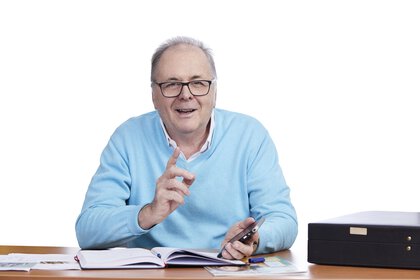 Mann im blauen Pullover sitzt lächelnd mit Stift in der Hand an einem Schreibtisch mit Papieren und einer Schachtel.
