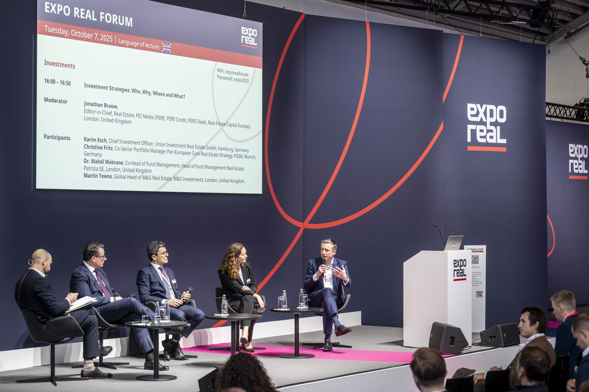A panel discussion at the EXPO REAL Forum: Five experts are seated on stage in front of a large presentation screen, discussing investment strategies. The speaker on the right is talking into a microphone while the others listen. Audience members sit in the foreground, and a lectern with the EXPO REAL logo stands on the right.