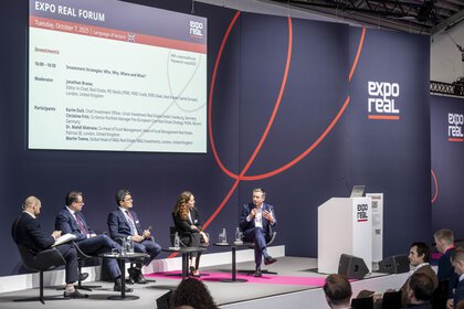 Eine Podiumsdiskussion beim EXPO REAL Forum: Fünf Fachleute sitzen auf einer Bühne vor einem großen Präsentationsbildschirm und diskutieren über Investmentstrategien. Ein Redner rechts spricht in ein Mikrofon, während die anderen aufmerksam zuhören. Im Vordergrund sitzt Publikum, rechts steht ein Rednerpult mit EXPO REAL Logo.