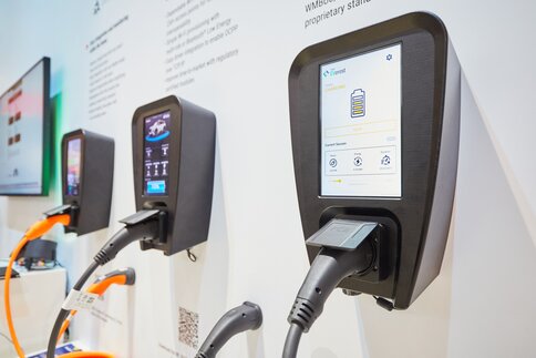 Three charging stations for electric vehicles with screens and plugged-in charging cables are displayed on one wall of an exhibition stand.