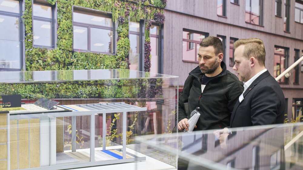 Zwei Männer betrachten aufmerksam ein detailliertes Architekturmodell eines modernen Gebäudes mit begrünten Fassaden und Holzverkleidung hinter Glas.