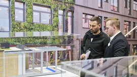 Zwei Männer betrachten aufmerksam ein detailliertes Architekturmodell eines modernen Gebäudes mit begrünten Fassaden und Holzverkleidung hinter Glas.