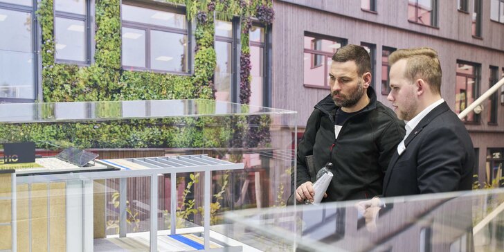Zwei Männer betrachten aufmerksam ein detailliertes Architekturmodell eines modernen Gebäudes mit begrünten Fassaden und Holzverkleidung hinter Glas.