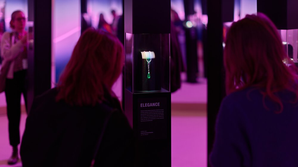 Menschen betrachten eine Halskette in einer schwach beleuchteten Ausstellung mit rosa und violettem Ambiente.