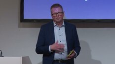 Ein Mann im Anzug hält ein Telefon und präsentiert vor einem Bildschirm.