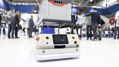 Ein niedriger Transportroboter, der in einer belebten Messehalle der productronica auf dem Boden steht. Er trägt eine graue Kiste.