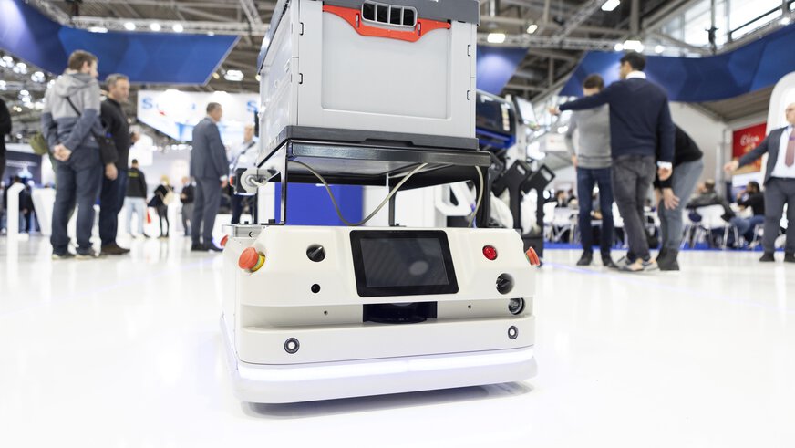Ein niedriger Transportroboter, der in einer belebten Messehalle der productronica auf dem Boden steht. Er trägt eine graue Kiste.