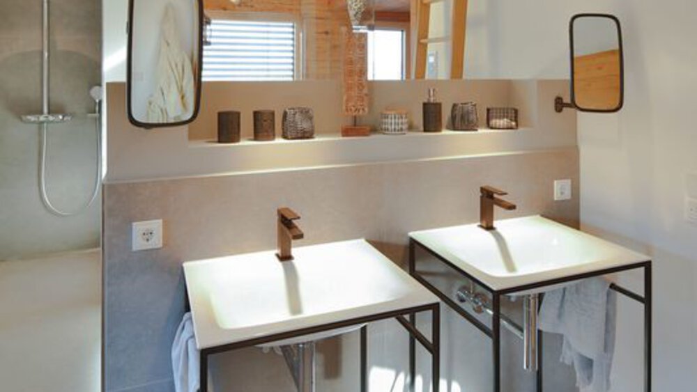 Modernes Badezimmer mit zwei weißen Waschbecken, wandmontierten Wasserhähnen und einem großen Spiegel darüber. Natürliches Licht durch ein Fenster.