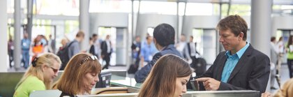 People at a busy check-in counter, with the trade fair entrance blurred in the background.