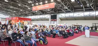 Lecture on the “travel stage” at the f.re.e trade fair with a large audience, red carpets and surrounding travel stands in an exhibition hall.