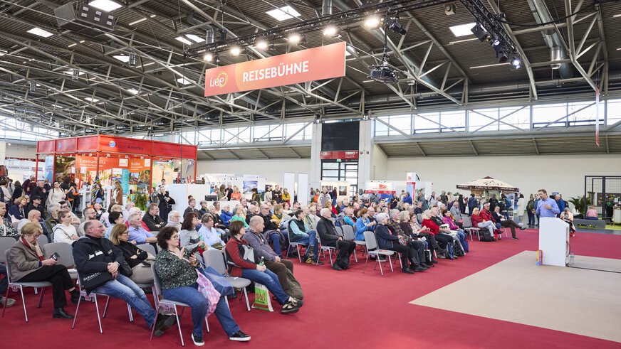 Lecture on the “travel stage” at the f.re.e trade fair with a large audience, red carpets and surrounding travel stands in an exhibition hall.