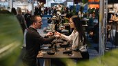 Eine Ausstellerin legt einem Messebesucher ein Armband ums Handgelenk. Im Hintergrund sind weitere Messestände und zahlreiche Besucher zu sehen.