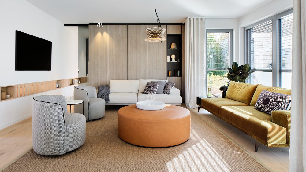 Modernes Wohnzimmer mit weißem Sofa, grauen Sesseln, großen Fenstern, einem Fernseher an der Wand und einer Pflanze.