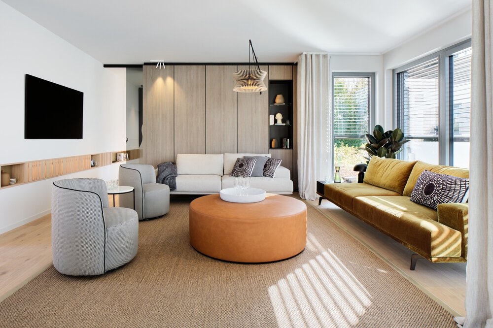 Modernes Wohnzimmer mit weißem Sofa, grauen Sesseln, großen Fenstern, einem Fernseher an der Wand und einer Pflanze.