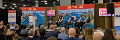 Panelists speak on stage to an audience at a conference with red banners and cityscapes in the background.