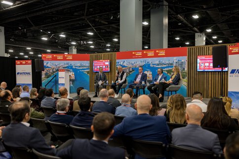 Panelists speak on stage to an audience at a conference with red banners and cityscapes in the background.