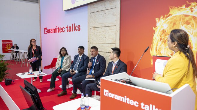 A person speaks at a podium to a panel of four seated and one standing person. A sign reads 