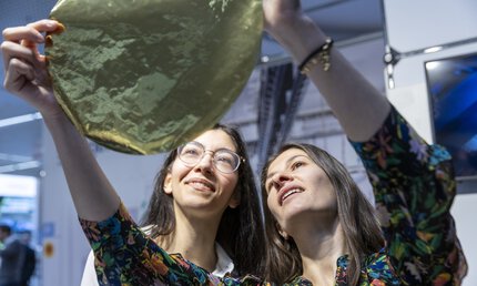 Two women are looking at a round, gold-coloured, flexible wafer at LOPEC.