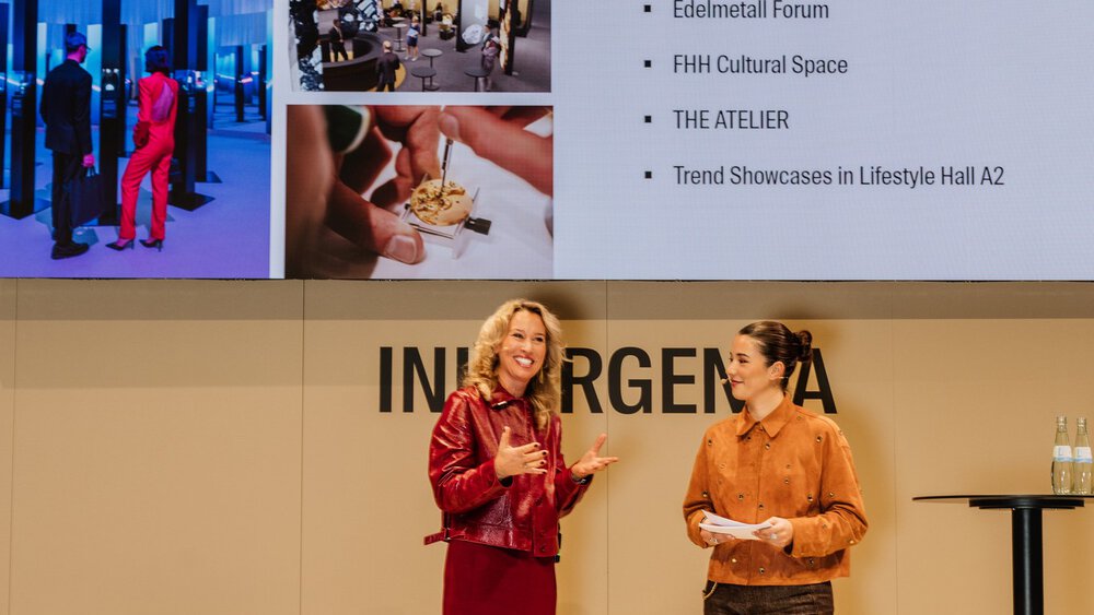 Two women are speaking on stage next to a screen showing highlights of the event and photos of previous activities.