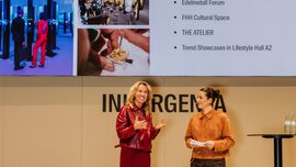 Two women are speaking on stage next to a screen showing highlights of the event and photos of previous activities.
