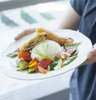 A person is holding a plate with rice, vegetables and a sliced spring roll.