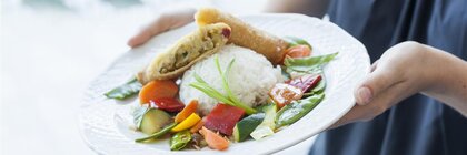A person is holding a plate in their hands on which rice, vegetables and spring rolls have been arranged attractively.