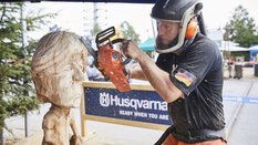 On the open-air grounds of Messe München, a man in protective clothing uses a small chainsaw to shape a caricature figure from a large piece of wood.
