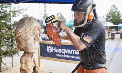 On the open-air grounds of Messe München, a man in protective clothing uses a small chainsaw to shape a caricature figure from a large piece of wood.