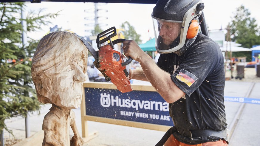 On the open-air grounds of Messe München, a man in protective clothing uses a small chainsaw to shape a caricature figure from a large piece of wood.